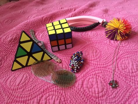 Alexis Simmerman has a number of stress toys and objects, including a pair of earmuffs, a ball of magnetic studs, two rubix cubes (one pyramid shaped) a necklace, a goo toy, a fuzzy ball, and a broken watch.