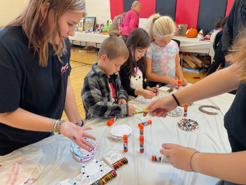 High school volunteers help elementary students create community show crafts utilizing a variety of materials such as glue sticks, templated paper, and crayons.